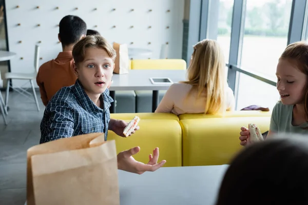 Surprised boy with sandwich gesturing while talking to classmates in dining room — Stock Photo