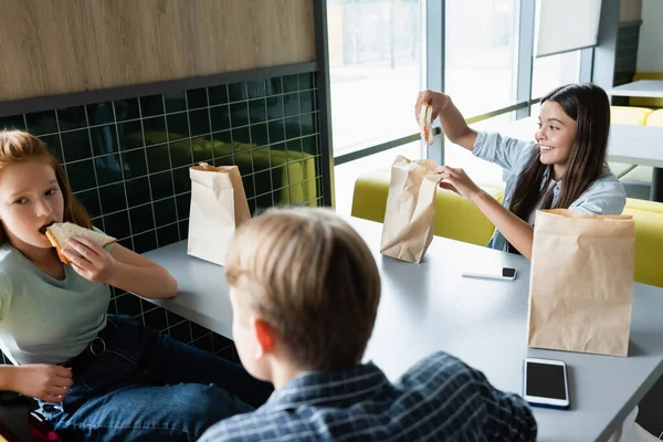 Feliz adolescente celebración sándwich cerca de compañeros de clase en la cantina de la escuela - foto de stock