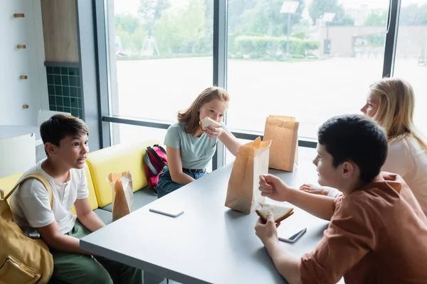 Multiethnic teenagers eating sandwiches while talking in school eatery during lunch break — Stock Photo
