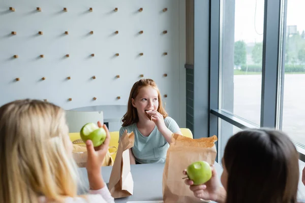 Chica pelirroja positiva comiendo sándwich cerca de compañeros de clase con manzanas en primer plano borrosa - foto de stock