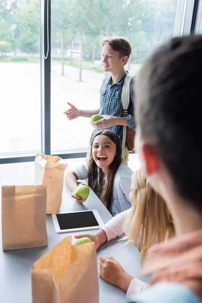 Excited girl holding apple while laughing near classmates in eatery — Stock Photo