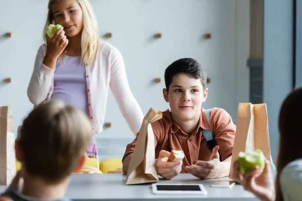 Asombrado adolescente gesto mientras habla a borrosa amigos en la escuela comedor - foto de stock