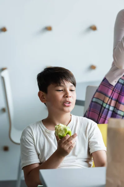 Asiatico ragazzo mangiare mela e indicando con medio dito mentre parlando in sala da pranzo — Foto stock