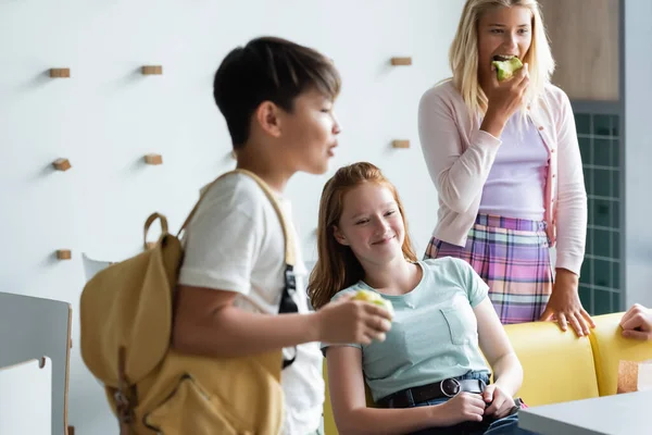 Allegra rossa ragazza seduta nella sala da pranzo della scuola vicino offuscata asiatico ragazzo e studentessa mangiare mela — Foto stock