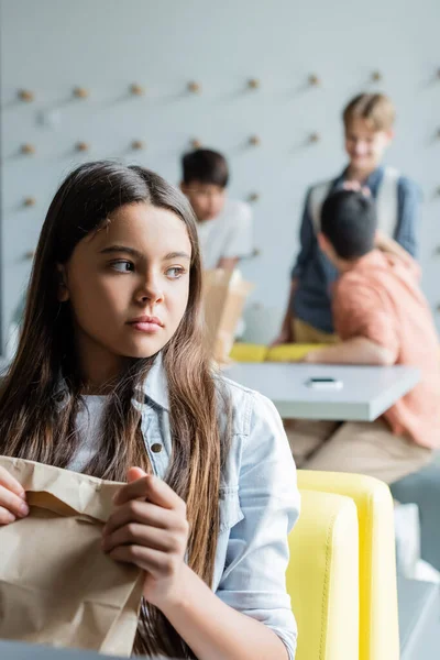 Menina frustrada sentado com saco de papel na sala de jantar perto de colegas de classe no fundo embaçado — Fotografia de Stock