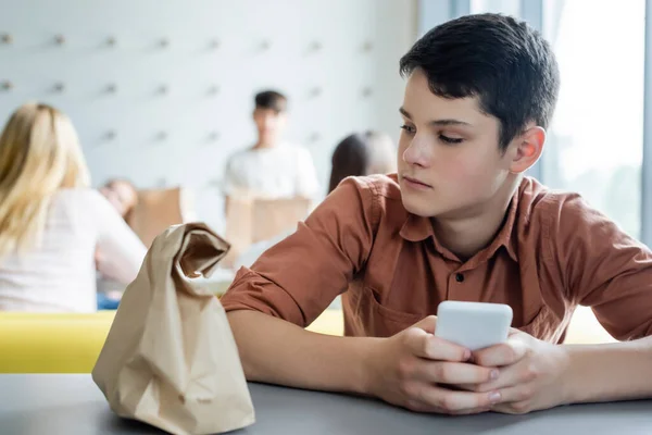 Сумний хлопчик з мобільним телефоном сидить один у шкільному харчуванні біля паперового мішка та розмитих однокласників — стокове фото
