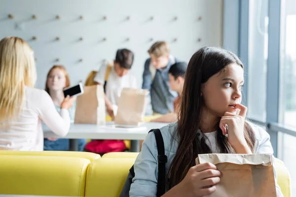 Depressed schoolgirl looking away while sitting with paper bag in canteen near blurred teenagers — Stock Photo