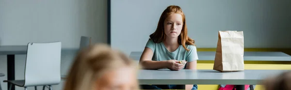 Triste ragazza con panino seduto da solo nella sala da pranzo della scuola in primo piano sfocato, striscione — Foto stock
