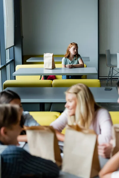 Upset schoolgirl sitting alone in dining room during lunch break near blurred teenagers — Stock Photo