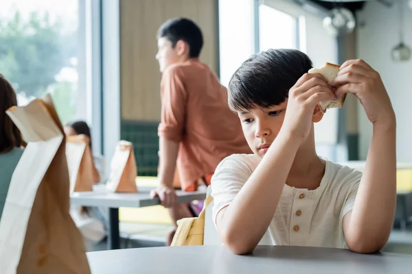 Sad asian boy holding sandwich while sitting alone in school canteen near blurred pupils — Stock Photo