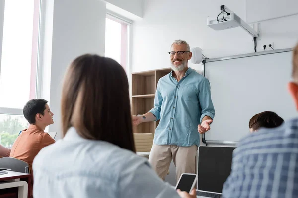 Positive teacher gesturing while speaking near teenagers during lesson in school — Stock Photo