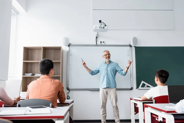 Вид сзади на одноклассников рядом с учителем, говорящих и показывающих пальцами во время урока — стоковое фото