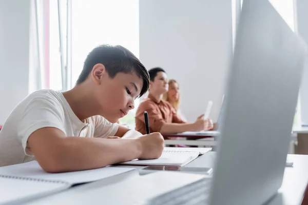 Asian schoolboy writing in notebook near blurred laptop and classmates — Stock Photo