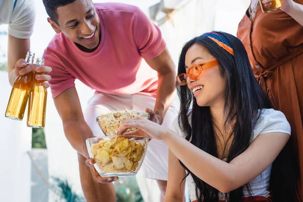 Feliz afro-americano homem propondo pipocas para asiático mulher durante festa com amigos — Fotografia de Stock