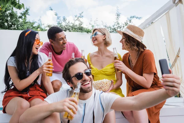 Homem alegre em óculos de sol tomando selfie perto de amigos inter-raciais beber cerveja — Fotografia de Stock