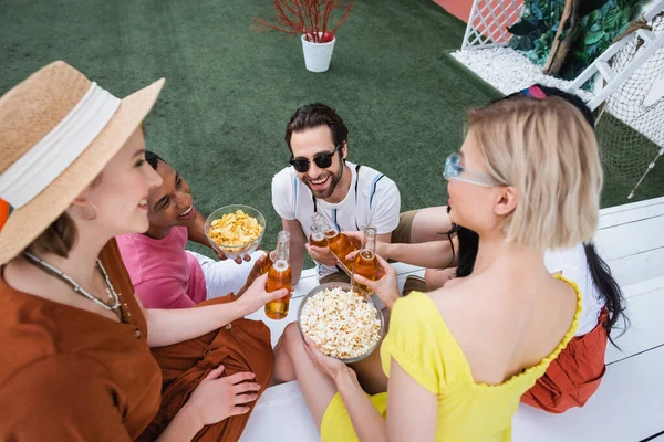 Overhead view of multiethnic friends clinking bottles of beer near chips and popcorn — Stock Photo