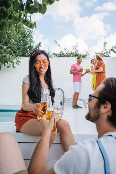 Азиатка в солнечных очках, звенящая бутылками пива с мужчиной рядом с мультикультурными друзьями на размытом фоне — стоковое фото