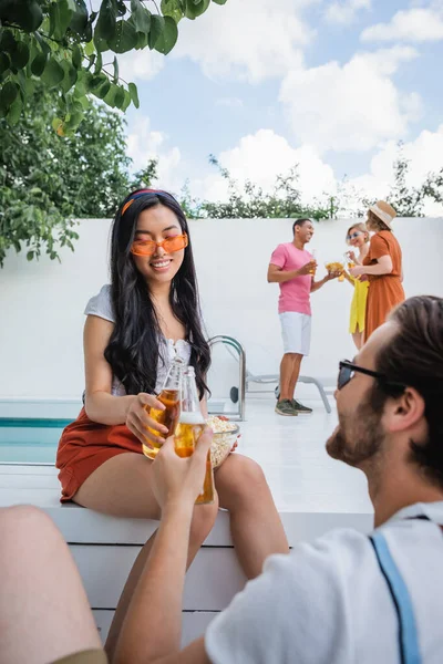 Asiatin mit Sonnenbrille klappert mit Mann in der Nähe multikultureller Freunde Bierflaschen auf verschwommenem Hintergrund — Stockfoto