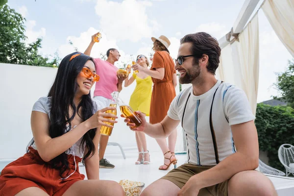 Happy asian woman clinking bottles of beer with man near cheerful multiethnic friends on background — Stock Photo