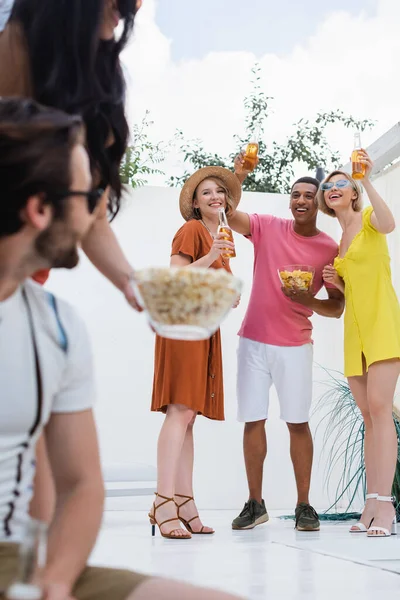 Cheerful multicultural friends toasting with beer near man and woman on blurred foreground — Stock Photo