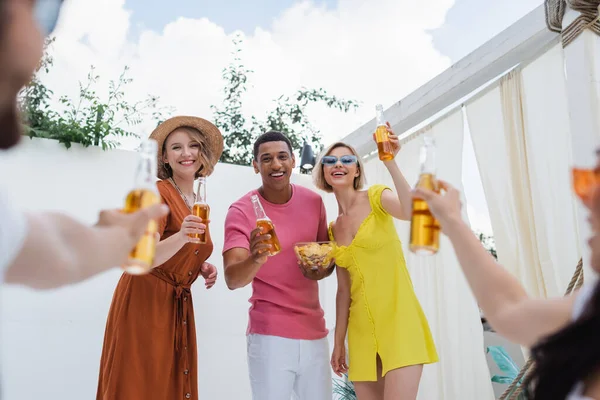 Возбужденные мультикультурные друзья тост с бутылками пива во время вечеринки на размытом переднем плане — стоковое фото