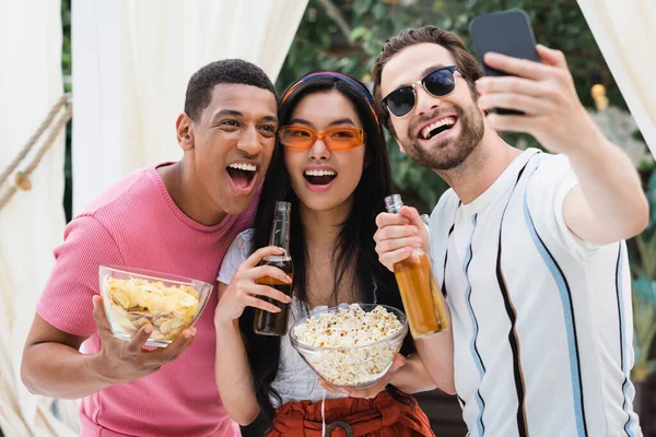 Excited multiethnic friends taking selfie with beer and snacks on cellphone — Stock Photo