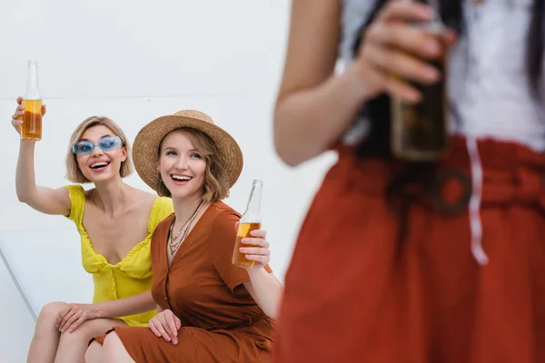 Cheerful women toasting with bottles of beer near blurred friend — Stock Photo