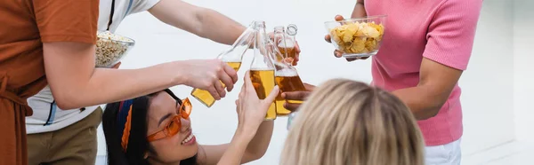 Amigos multiétnicos tintineo botellas de cerveza cerca de patatas fritas y palomitas de maíz durante la fiesta, pancarta - foto de stock