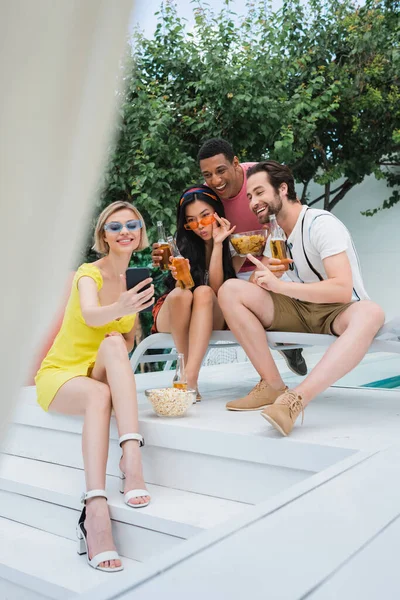 Femme heureuse prenant selfie sur téléphone portable pendant la fête avec des amis multiethniques, premier plan flou — Photo de stock