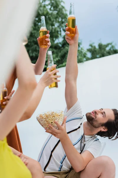 Fröhlicher Mann mit Popcorn, der mit Bier neben Frauen im verschwommenen Vordergrund anstößt — Stockfoto