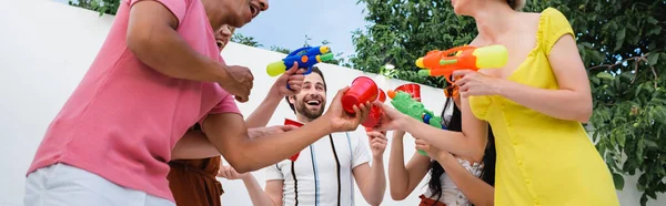 Excited multicultural friends with water pistols toasting with plastic cups during party, banner — Stock Photo