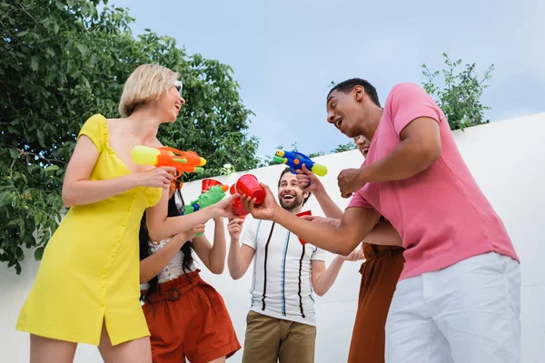 Alegres amigos multiculturales brindando con copas de plástico mientras sostiene pistolas de agua - foto de stock