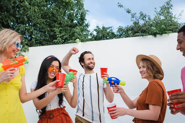 Cheerful man showing win gesture near multicultural friends with water pistols and plastic cups — Stock Photo