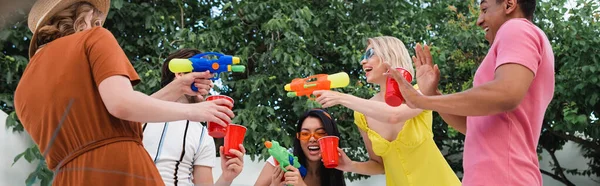 Aufgeregte Frauen, die mit Wasserpistolen beim Sommerfest mit multiethnischen Freunden spielen, Banner — Stockfoto