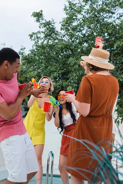 Excited multiethnic friends having fun with water pistols during summer party — Stock Photo