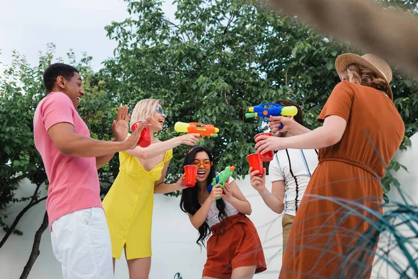 Fröhliche Frauen spielen mit Wasserpistolen in der Nähe lachender multiethnischer Freunde — Stockfoto