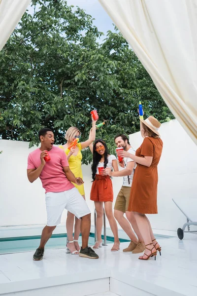 Cheerful multiethnic friends with plastic cups and water pistols having fun in patio at poolside — Stock Photo