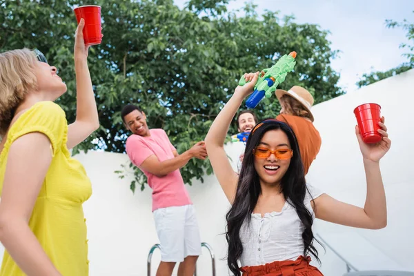 Alegre asiático mujer celebración de plástico taza y agua pistola durante partido con multiétnicos amigos - foto de stock