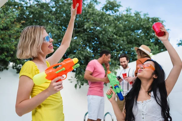 Excited interracial women toasting with plastic cups while holding water pistols — Stock Photo
