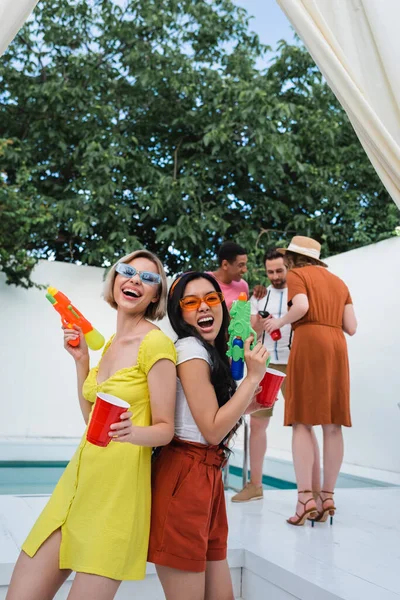 Interracial women with water pistols laughing while standing back to back near multiethnic friends — Stock Photo