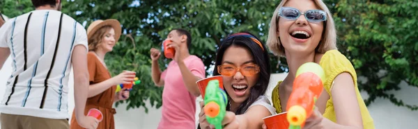 Fröhliche interrassische Frauen mit Wasserpistolen, die lachen, während sie in die Kamera neben verschwommenen Freunden schauen, Banner — Stockfoto