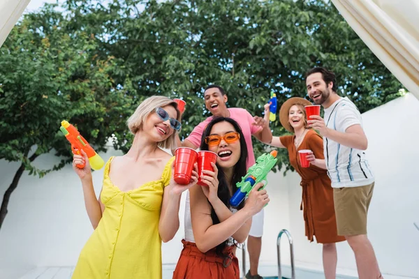 Mujeres interracial con pistolas de agua riendo y tostando con vasos de plástico cerca de amigos felices - foto de stock