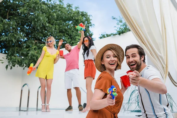 Laughing man pointing at camera near woman in straw hat and blurred multiethnic friends with water pistols — Stock Photo