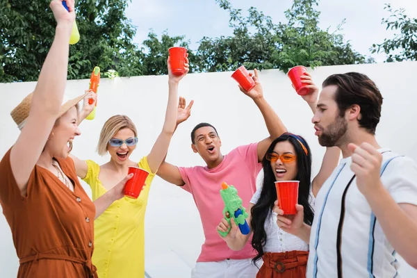 Amigos multiétnicos llenos de alegría con vasos de plástico y pistolas de agua que muestran el gesto de ganar durante la fiesta - foto de stock