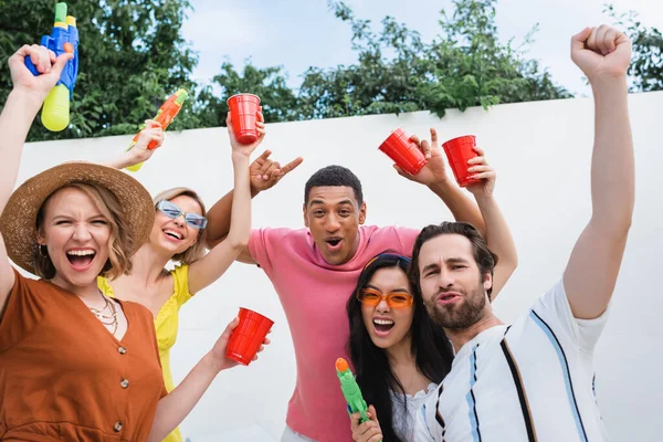 Amigos multiétnicos emocionados con copas de plástico y pistolas de agua mostrando rock y ganar gestos durante la fiesta - foto de stock