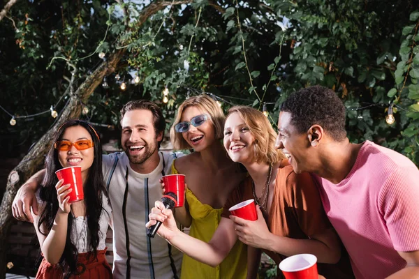 Amigos multiétnicos felices con copas de plástico cantando karaoke en el jardín nocturno - foto de stock