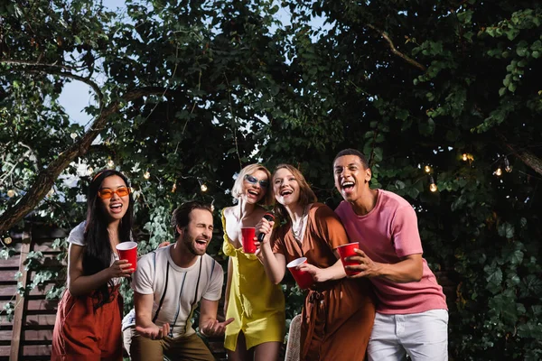 Amigos multiculturais com bebidas cantando karaoke ao ar livre durante a festa — Fotografia de Stock