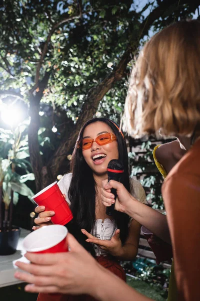 Азійська жінка з пластиковою чашкою співає біля розмитих друзів з мікрофоном на вулиці. — стокове фото