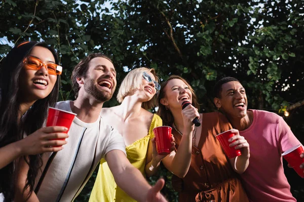 Gente alegre y multiétnica cantando karaoke y bebiendo en vasos de plástico al aire libre - foto de stock