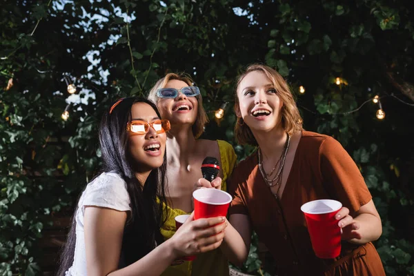 Donne interrazziali con coppe di plastica che tengono il microfono all'aperto — Foto stock